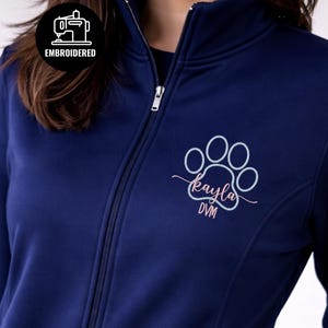 May include: Navy blue zip-up jacket with a high collar. Features an embroidered paw print design with the name "Kayla DVM" in pink and light blue. The jacket has a "EMBROIDERED" patch.