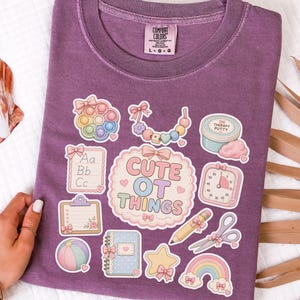 May include: A purple Comfort Colors t-shirt with various cute OT (Occupational Therapy) themed graphics. The graphics include a pop-it toy, a rainbow, a notepad, and the text "Cute OT Things".