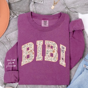 May include: A purple sweatshirt with the name "BIBI" in a floral design. The sleeve has the names "amelia", "john", and "jennifer" printed on it. The sweatshirt is displayed with a gray jacket, blue jeans, and a brown hat.