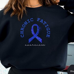 Könnte beinhalten: Schwarzes Sweatshirt mit einer blauen Schleifengrafik und den Worten "CHRONIC FATIGUE" in einem kreisförmigen Design. Unter dem Band steht das Wort "awareness". Das Sweatshirt ist einfarbig schwarz.