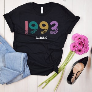 May include: Black t-shirt with the year 1993 printed in colorful letters and the word "Classic" underneath. The shirt is displayed on a white wooden surface with a pair of blue jeans, a bouquet of pink flowers, and black high heels.