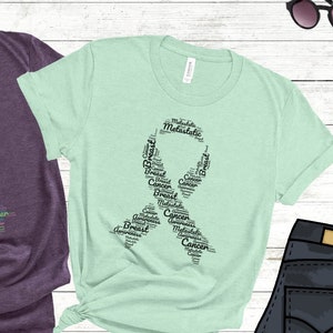 K&ouml;nnte beinhalten: Zwei T-Shirts mit einem rosa und t&uuml;rkisfarbenen Band aus W&ouml;rtern, die mit Brustkrebs zusammenh&auml;ngen. Die Shirts sind in den Farben Heather Purple und Heather Mint.