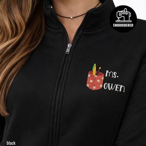 Può includere: Giacca nera con zip e tasca ricamata. La tasca presenta un motivo floreale rosso e contiene penne e matite. Il nome "ms. OWEN" è ricamato sotto. La giacca ha un collo alto e una chiusura con cerniera.