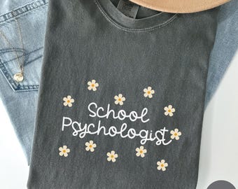 May include: A dark grey t-shirt with the words "School Psychologist" in white script, decorated with small daisy graphics. The shirt is folded on blue jeans, with a tan hat and a silver necklace visible.