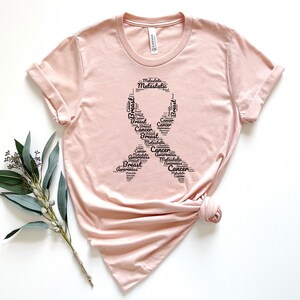 K&ouml;nnte beinhalten: Ein pinkfarbenes T-Shirt mit einem schwarzen Banddesign, das aus W&ouml;rtern besteht, die mit der Sensibilisierung f&uuml;r Brustkrebs zusammenh&auml;ngen. Die W&ouml;rter umfassen "Breast", "Cancer", "Awareness", "Metastatic" und "Ribbon".