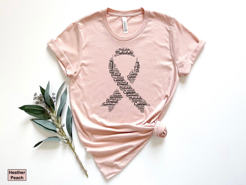 K&ouml;nnte beinhalten: Ein pinkfarbenes T-Shirt mit einem schwarzen Banddesign, das aus W&ouml;rtern besteht, die mit der Sensibilisierung f&uuml;r Brustkrebs zusammenh&auml;ngen. Die W&ouml;rter umfassen "Breast", "Cancer", "Awareness", "Metastatic" und "Ribbon".
