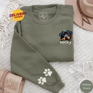 Peut inclure: Un sweat-shirt vert avec un Rottweiler qui sort de la poche. Le nom du chien est "Rocky" et est imprimé sous l'image. Le sweat-shirt a des empreintes de pattes blanches sur les manches.