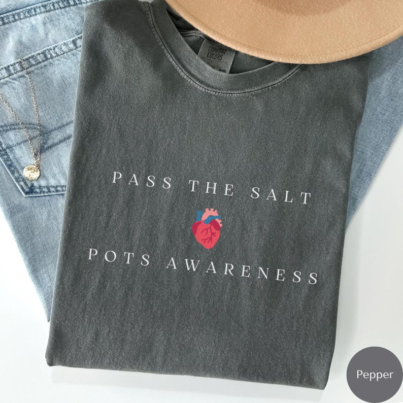 Op de afbeelding: Een grijs T-shirt met de tekst "Pass the Salt" en een rode hartgrafiek. Onder de tekst staat "POTS Awareness".