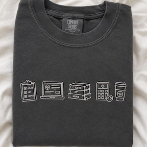 Puede incluir: Una camiseta gris carbón de cuello redondo con un gráfico de líneas blancas que muestra un portapapeles con casillas de verificación ICD-10, CPT y HCPCS, un portátil, tres libros etiquetados ICD-10, CPT y HCPCS, una calculadora y un vaso de café.