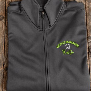 Könnte beinhalten: Eine anthrazitfarbene Reißverschlussjacke mit einer Stickerei auf der Brust. Die Stickerei zeigt den Text "OFFICE MANAGER" in Limettengrün, ein weißes Zahnsymbol und den Namen "Kaitie" in limettengrüner Schreibschrift.
