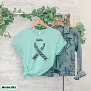 K&ouml;nnte beinhalten: Ein T-Shirt in der Farbe Mint mit einem schwarzen Banddesign, das aus W&ouml;rtern besteht, die mit dem Bewusstsein f&uuml;r Brustkrebs zusammenh&auml;ngen. Die W&ouml;rter beinhalten "Breast", "Cancer", "Awareness", "Metastatic" und "Multifocal".
