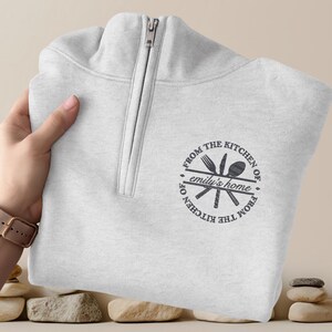 Può includere: Una felpa grigio chiaro con zip a un quarto e cerniera argentata. La felpa presenta una grafica circolare con forchetta, coltello e cucchiaio e il testo "FROM THE KITCHEN OF emily's home FROM THE KITCHEN OF".