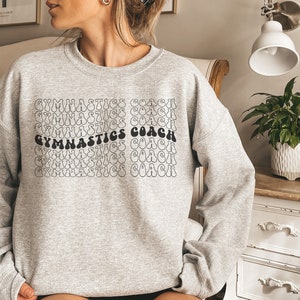 May include: Gray sweatshirt with the text "GYMNASTICS COACH" repeated in a retro, wavy font.