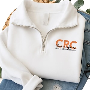 May include: White quarter-zip sweatshirt with a silver zipper. The sweatshirt features the text "CRC Jennifer Clinical Research Coordinate" in orange and black. The sweatshirt is folded on top of blue jeans.