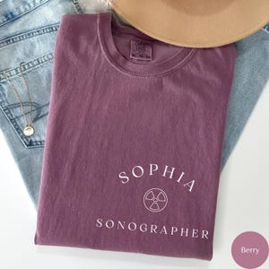 May include: A purple t-shirt with the text "Sophia Sonographer" and a white graphic of a medical symbol.