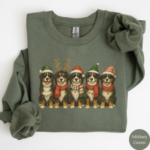 May include: Military green sweatshirt featuring a graphic of five Bernese Mountain Dogs wearing Christmas hats, scarves, and sunglasses. Each dog is adorned with festive lights. The dogs are arranged in a row, creating a holiday theme.