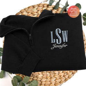 May include: Black quarter-zip sweatshirt with embroidered initials "LSW" and the name "Jennifer" in a light blue script. The sweatshirt is folded on a woven basket, with green sprigs around it. A sewing machine logo is in the top right corner.