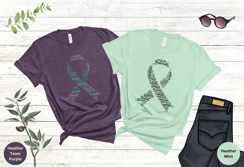 K&ouml;nnte beinhalten: Zwei T-Shirts mit einem rosa und t&uuml;rkisfarbenen Band aus W&ouml;rtern, die mit Brustkrebs zusammenh&auml;ngen. Die Shirts sind in den Farben Heather Purple und Heather Mint.