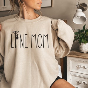 May include: A beige sweatshirt with a black graphic of a lineman on a power pole and the text "LINE MOM".