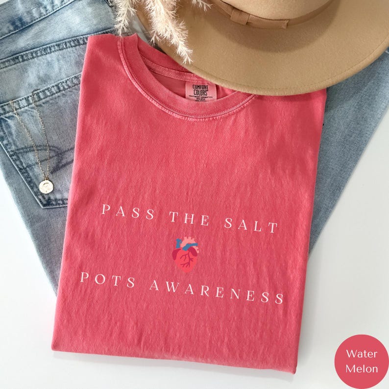 Op de afbeelding: Een rood T-shirt met de tekst "Pass the Salt" en een rood hart met een blauwe omtrek. Onder het hart staat de tekst "POTS Awareness". Het shirt is gevouwen en ligt op een blauwe spijkerbroek.