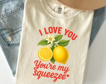 Könnte beinhalten: Cremefarbenes T-Shirt mit dem roten Text "I Love You, You're my Squeezee". Das Shirt zeigt eine Zitronengrafik mit einer Zitronenscheibe, einer ganzen Zitrone, Blättern und Blüten. Das Shirt liegt auf einer Jeans.