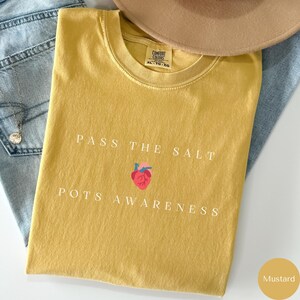 Op de afbeelding: Mosterdgeel T-shirt met de tekst "Pass the Salt POTS Awareness" en een gestileerd rood hart met een gestileerd anatomisch hart erin.