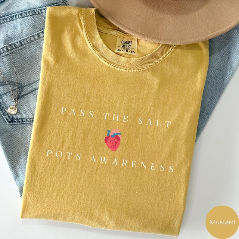 Op de afbeelding: Mosterdgeel T-shirt met de tekst "Pass the Salt POTS Awareness" en een gestileerd rood hart met een gestileerd anatomisch hart erin.