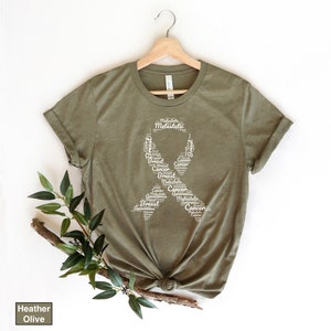 K&ouml;nnte beinhalten: Ein T-Shirt in der Farbe Heide-Olive mit einem wei&szlig;en Band-Design aus W&ouml;rtern, die mit dem Bewusstsein f&uuml;r Brustkrebs zusammenh&auml;ngen. Die W&ouml;rter beinhalten "Brust", "Krebs", "Bewusstsein", "metastasiert" und "Metastasen".
