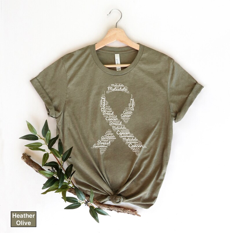 K&ouml;nnte beinhalten: Ein T-Shirt in der Farbe Heide-Olive mit einem wei&szlig;en Band-Design aus W&ouml;rtern, die mit dem Bewusstsein f&uuml;r Brustkrebs zusammenh&auml;ngen. Die W&ouml;rter beinhalten "Brust", "Krebs", "Bewusstsein", "metastasiert" und "Metastasen".