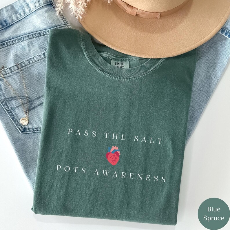 Op de afbeelding: Een groen T-shirt met de tekst "Pass the Salt POTS Awareness" en een rood hart met een gestileerd anatomisch hartontwerp.