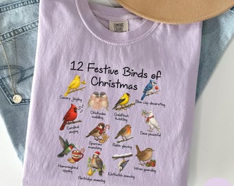 May include: Orchid-colored t-shirt with the text "12 Festive Birds of Christmas" and illustrations of various birds. The shirt features a crew neck and is made of a soft material. The Comfort Colors brand is visible on the shirt.