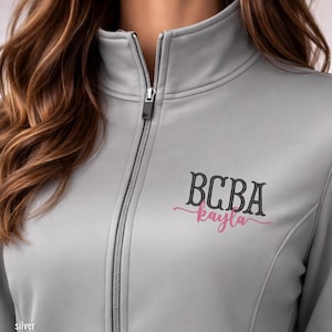 May include: A silver-colored zip-up jacket with a high collar. The jacket features the embroidered text "BCBA Kayla" in black and pink script on the chest. The zipper is silver.