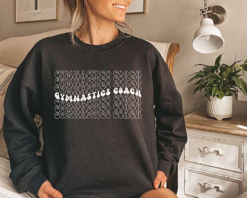 May include: Black sweatshirt with white text that reads "Gymnastics Coach" repeated in a retro font.