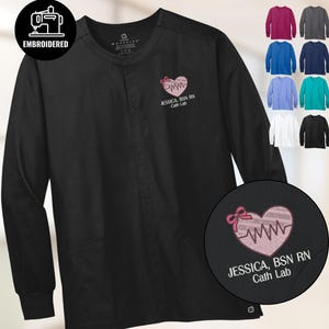 May include: Black long-sleeve medical scrub jacket with embroidered heart design and text "JESSICA, BSN RN Cath Lab". Additional color options are displayed. The jacket features a round neck and button closure.