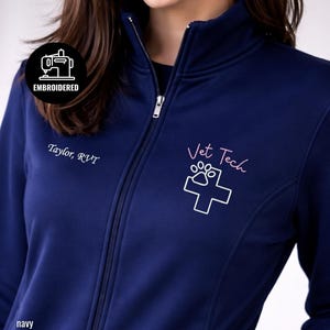 May include: Navy blue zip-up jacket with embroidered text and graphics. Features the words "Vet Tech" in pink script, a paw print and cross design, and the name "Taylor, RVT". The jacket also has an "EMBROIDERED" patch.