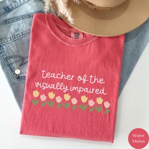 May include: A coral-colored t-shirt with the text "teacher of the visually impaired" in white script. Below the text is a row of yellow and pink tulips with green stems and leaves. A tan hat and blue jeans are in the background.