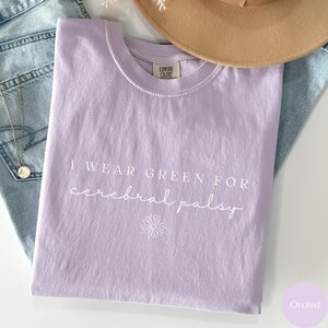 May include: A lavender t-shirt with the text "I wear green for cerebral palsy" in white lettering and a small white daisy graphic. The shirt is laid flat on a blue denim background.