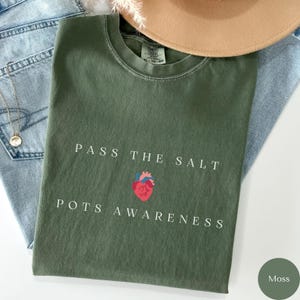 Op de afbeelding: Een groen T-shirt met de tekst "Pass the Salt Pots Awareness" en een rode hartgrafiek. Het shirt ligt op een blauwe denim achtergrond.