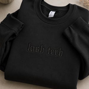 Peut inclure: Un sweat-shirt noir à col rond avec les mots "lash tech" brodés en noir dans une police de style gothique au centre de la poitrine. Le vêtement présente des manches longues avec des poignets côtelés et un col côtelé, avec une étiquette blanche.