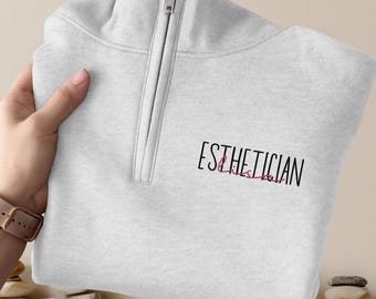 May include: A light gray quarter-zip sweatshirt with the word "ESTHETICIAN" printed on the front in black script with the word "Loves" in pink script. The sweatshirt has a silver zipper and a folded collar.
