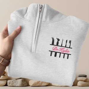 May include: A light gray quarter-zip sweatshirt with a black and pink embroidered design. The design features dental tools and the text "Dr. Kylie". The sweatshirt has a silver zipper.
