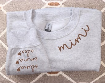 May include: A pale grey sweatshirt with the word "mimi" in brown script on the front. The sleeve displays the names "olivia", "emma", and "alex" in brown script, each with a small heart symbol.