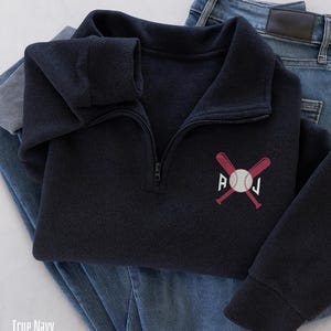 Peut inclure: Un pull-over bleu marine avec une encolure zippée, orné d'une broderie de baseball avec des battes croisées et les initiales "A O J". Le pull est présenté sur un jean bleu.