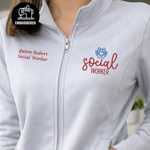 May include: Silver zip-up jacket with embroidered text and graphics. The jacket features the words "Quinn Robert Social Worker" in red script on the left chest. The right chest displays the words "social worker" in red script with a blue graphic above it. The jacket has a silver zipper.