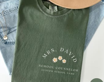 May include: A green t-shirt with the text "Mrs. David School Counselor Custom School Name" and three white daisies with orange centers.