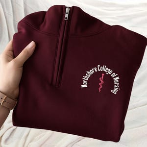 May include: A maroon quarter-zip sweatshirt with the text "Northshore College of Nursing" in a white, arched font. A pink caduceus symbol is centered below the text. The sweatshirt has a silver zipper.