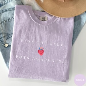 Op de afbeelding: Een lavendelkleurig T-shirt met de tekst "Pass the Salt" en een gestileerd hart met de tekst "POTS Awareness" eronder.