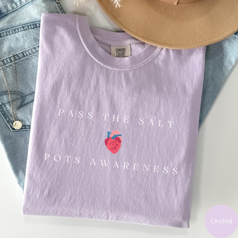 Op de afbeelding: Een lavendelkleurig T-shirt met de tekst "Pass the Salt" en een gestileerd hart met de tekst "POTS Awareness" eronder.