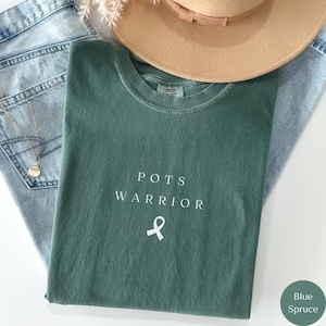 May include: A green t-shirt with the text "POTS WARRIOR" and a white ribbon printed on the front. The shirt is folded and laid on top of a pair of blue jeans.
