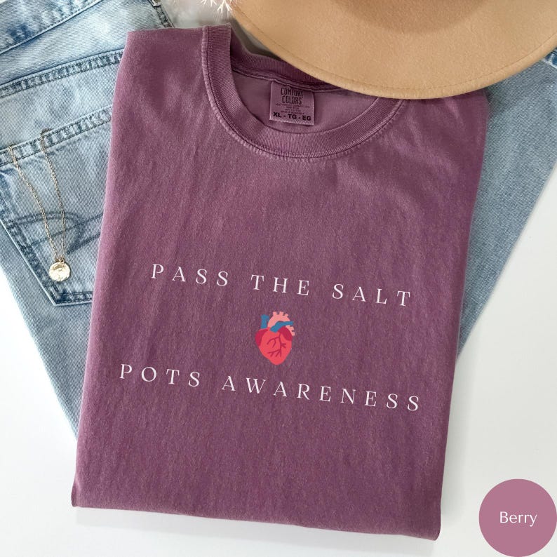 Op de afbeelding: Een paars T-shirt met de tekst "Pass the Salt POTS Awareness" en een gestileerde rode hartgrafiek.